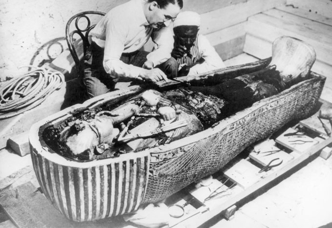 black and white photo of Howard Carter and an Egyptian member of his team kneel beside Tutankhamun's sarcophagus, the lid of which has been removed