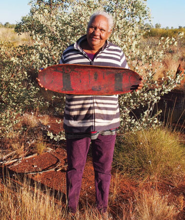 A photograph of Anna Rivers holding a long, red boat with black boab trees painted on the surface