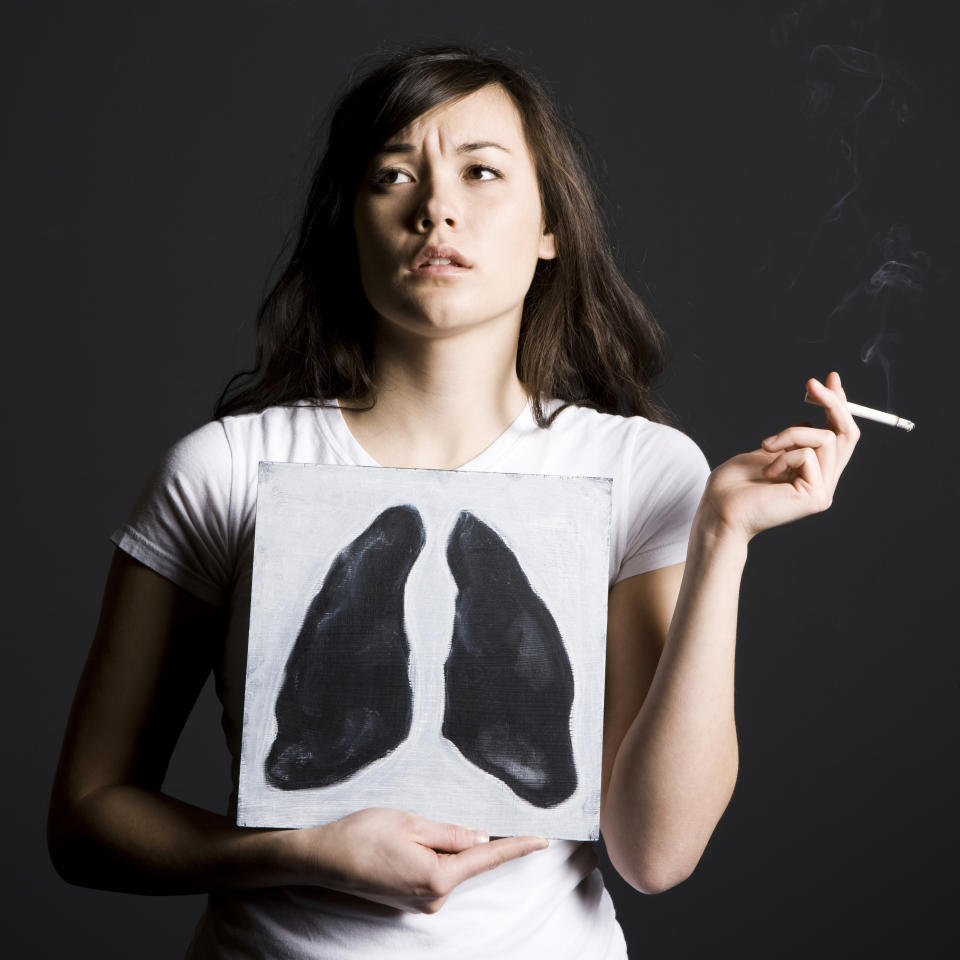 Woman smoking on a black background holding a poster with black lungs on it.