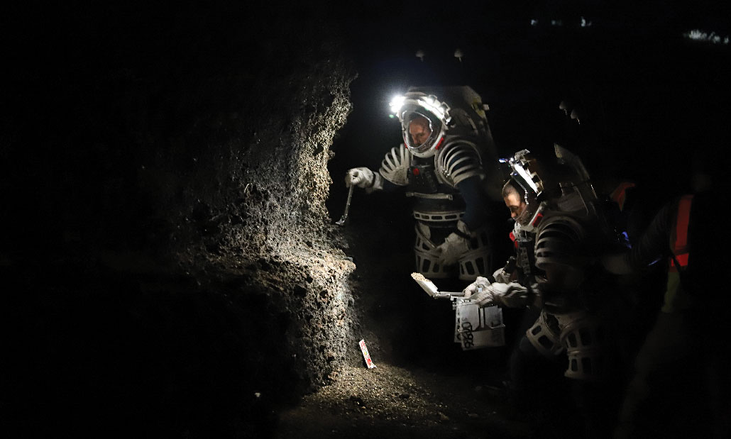 NASA engineers wear spacewalk backpacks with headlamps in a dark space