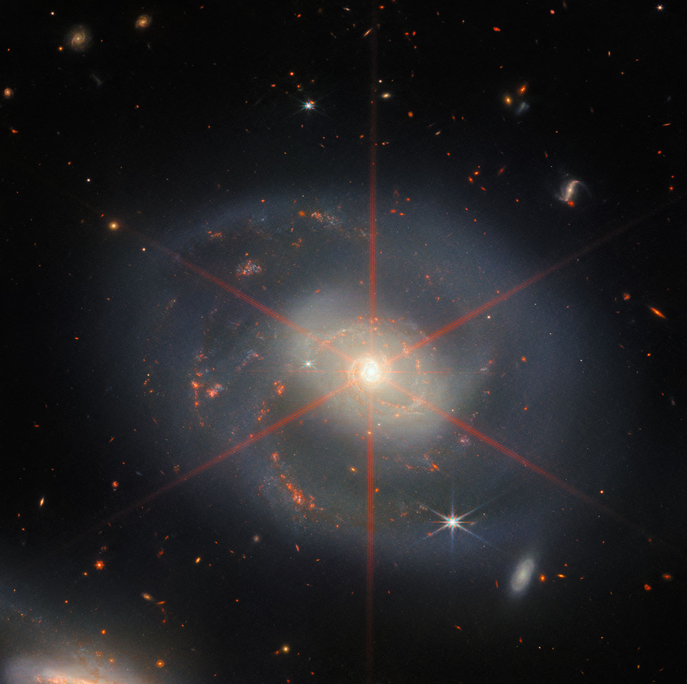 This image shows the spiral galaxy that dominates the bright central region.  The galaxy has blue-purple regions with orange regions filled with stars.  There is also a large visible diffraction peak that appears like a star in the central region of the galaxy.  Lots of stars and galaxies fill the scene